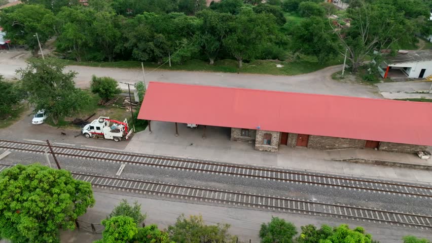 Rural train station with red roof surrounded by greenery and railway tracks in aerial orbital shot