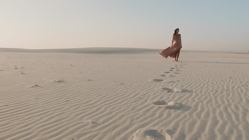 A graceful woman in a flowing pink dress walks across a vast desert, leaving a trail of footprints in the soft sand. The scene exudes calm, elegance, and solitude, perfect for conceptual visuals.
