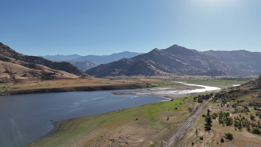 A scenic drive toward the majestic sequoias, featuring a stunning lake view surrounded by lush greenery and distant mountains. The calm water mirrors the clear blue sky, offering a tranquil vibe.