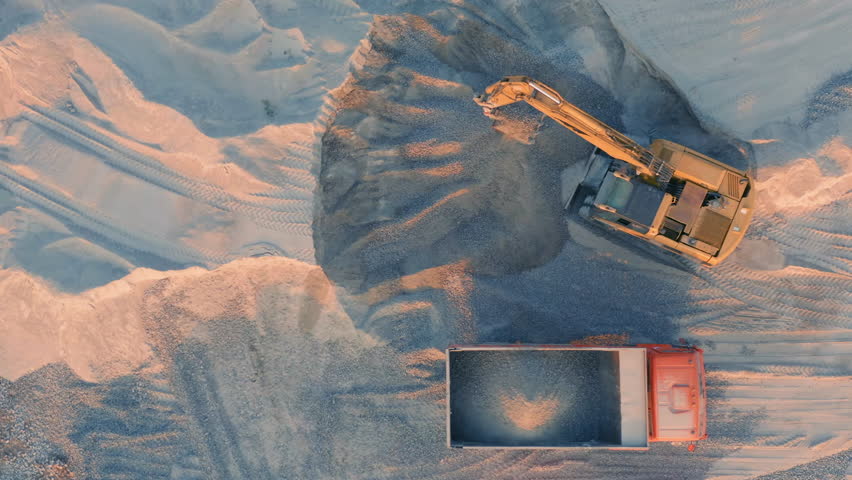 Heavy excavator loading crushed stone into orange dump truck in quarry at golden sunset light, aerial top down view of mining site