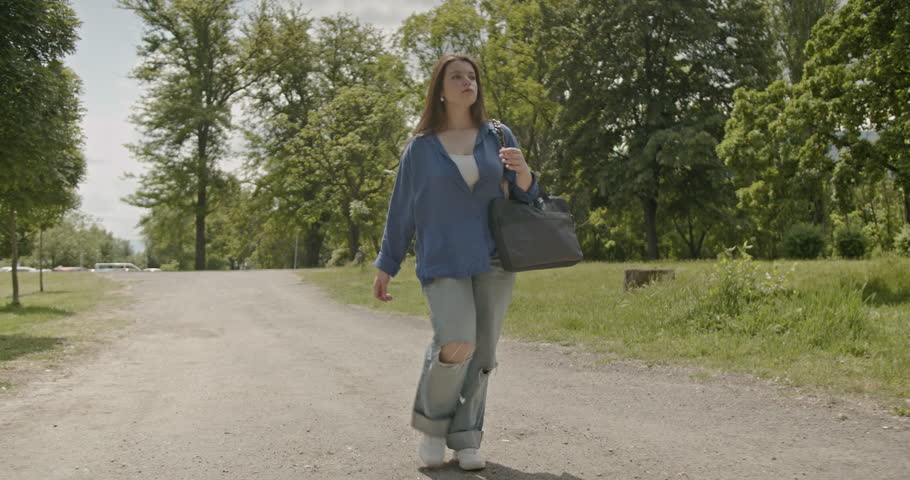 A young woman walks down a path while looking at her watch and waving down a car. She is carrying a bag over her shoulder.