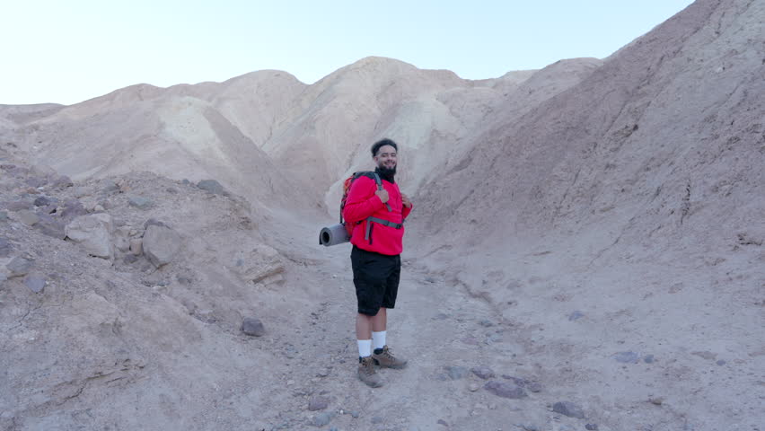 A cheerful tourist with a red backpack enjoys a hike in Death Valley National Park, surrounded by stunning rock formations and expansive desert views that stretch into the horizon