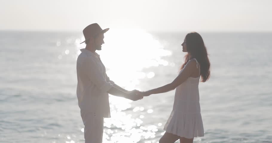 A romantic couple dances by the seaside during sunset, dressed in light summer clothing. The tranquil ocean and glowing sunlight create a dreamy, intimate atmosphere perfect for concepts of love and