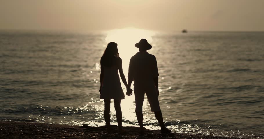 A romantic couple stands hand-in-hand on the beach, gazing at the golden sunset over the calm ocean. Their silhouette against the glowing water symbolizes peace, love, and togetherness.