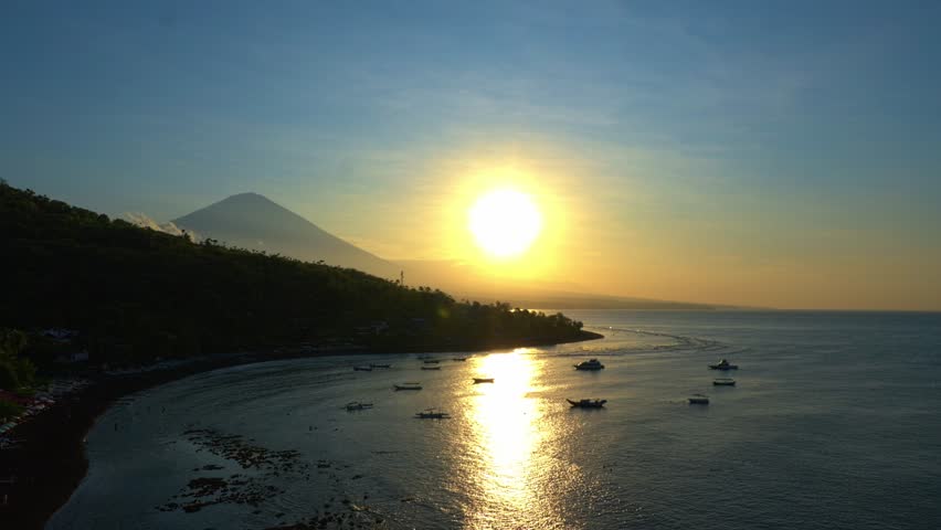 A serene sunset over a calm sea, with boats anchored near the shore. The sun casts a golden reflection on the water, while a mountain looms in the background, surrounded by lush greenery.