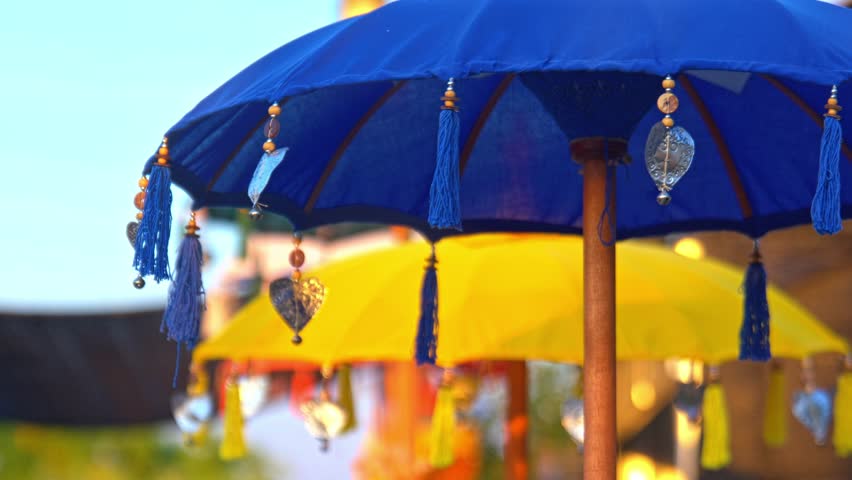 Colorful decorative umbrellas with tassels and ornaments, set against a bright blue sky. The foreground features a blue umbrella, while a yellow one is blurred in the background, creating a vibrant