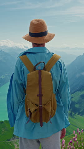 Vertical video. Handsome teenage boy with hiking backpack traveling in Switzerland standing on top of a mountain above the beautiful lake Interlaken in the background beautiful alpine mountains