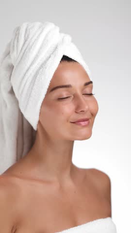 Portrait of a serene brunette woman with a towel turban, gently touching her face and smiling, showcasing glowing skin. Perfect for skincare, wellness, or beauty product promotions. 