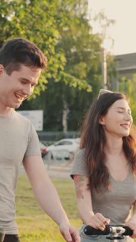 Friends communicating with each other while walking. Pretty asian girl and handsome guy in front of orange sunset passing by architectural building or park with a board and scooter in hands.