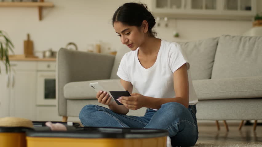 Hindu woman Indian girl Arabian female at home prepare for trip vacation travel using browsing smartphone order fly chat mobile phone booking hotel packing clothes in suitcase holding tickets passport