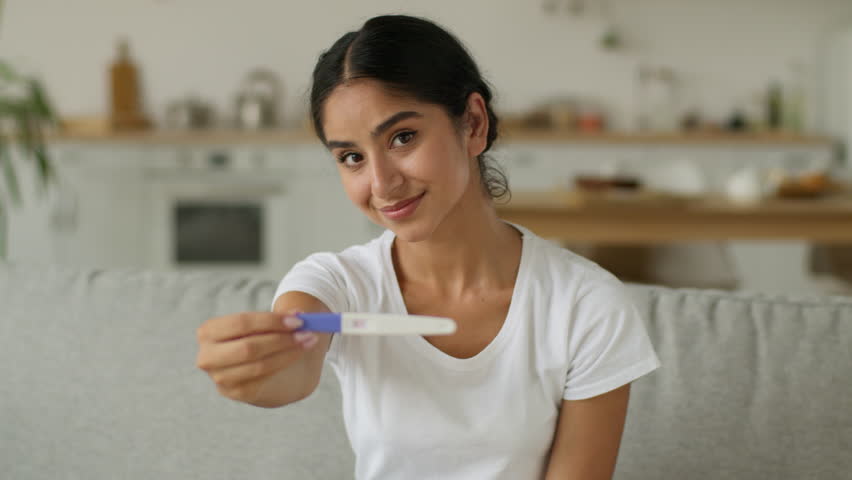 Health medicine happy healthy carefree Hindu pregnant woman Indian girl Arabian female at home with positive pregnancy test showing at camera smiling child anticipation childbirth motherhood family
