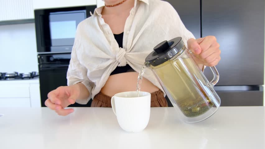 A woman makes a cup of herbal tea in her kitchen. tea steeps for few minutes and then is poured into cup. The woman enjoys the delicious and aromatic beverage.