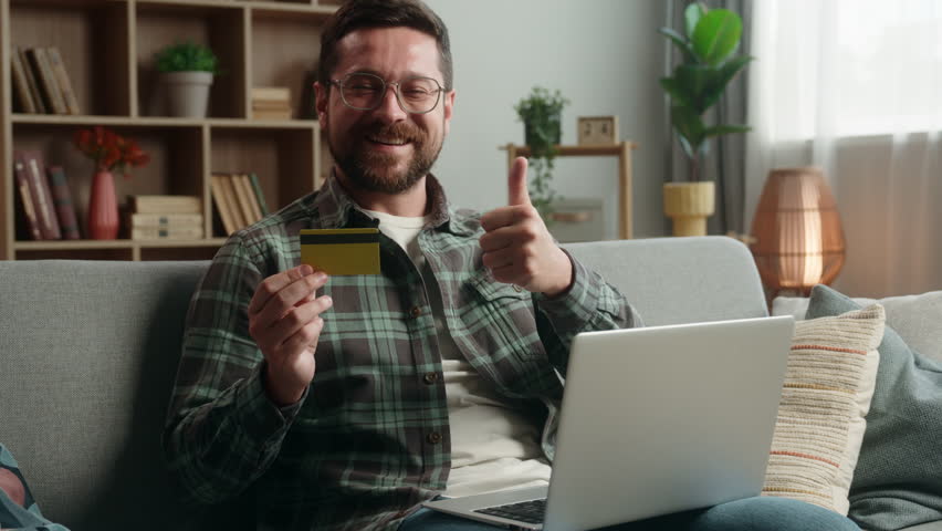 Caucasian man male guy at home happy smiling holding credit bank card buying online internet banking pay purchase shopping buying payment success achieve with laptop computer showing thumb up order