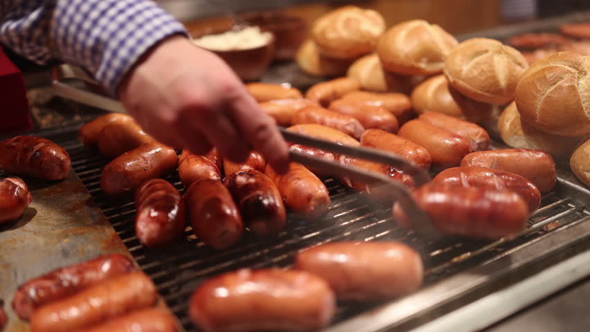 A detailed closeup of succulent and juicy sausages grilling to perfection on a hot barbecue, with warm, fluffy buns in the background, capturing the lively and vibrant atmosphere of a food festival
