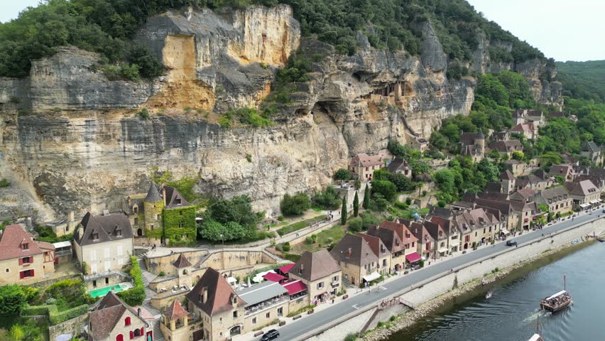 Drone aerial view in France countryside small old medieval town below a stone cliff surrounded by a green forest and a river circling around in La Roque Gageac