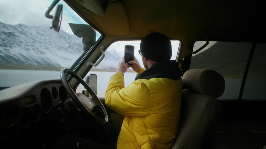 Handsome man wearing a bright yellow jacket is photographing on phone the stunning mountain scenery from inside a vehicle