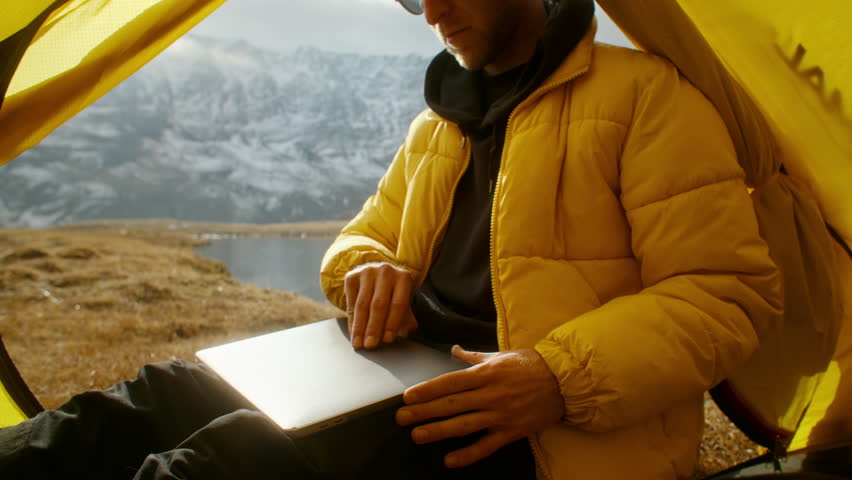 A person sits on yellow tent working diligently on a laptop while surrounded by breathtaking mountain scenery and landscape views