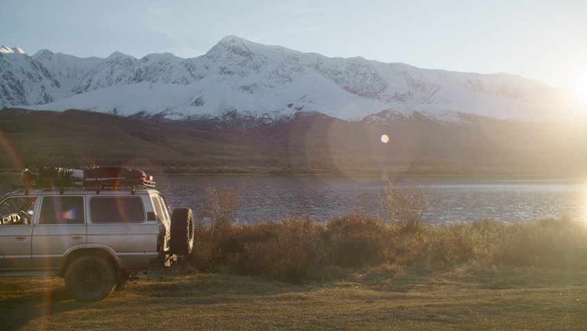 Young Man climbs onto the roof of car and looks into the distance. SUV is parked close to serenelake, surrounded by majestic mountain scenery