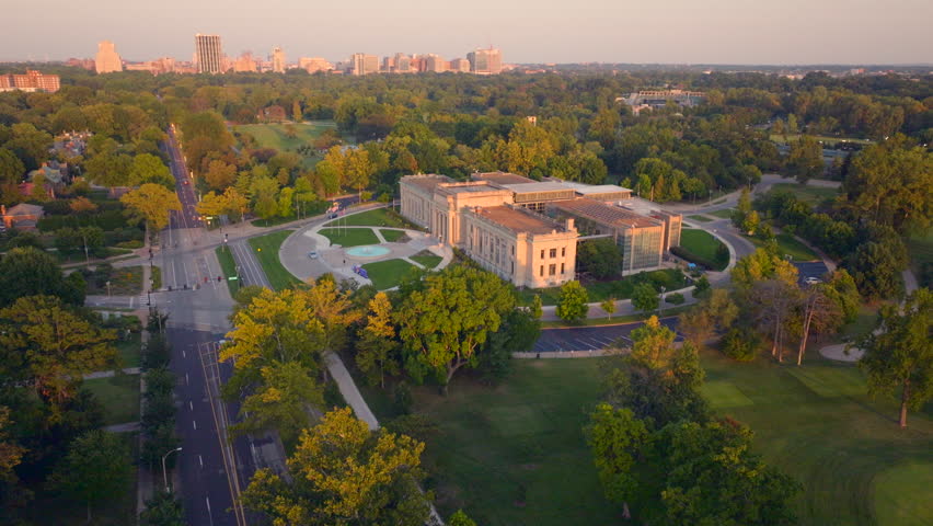 Aerial over Lindell Blvd towards the Missouri History Museum in Forest Park in St. Louis, Missouri at golden hour in summer with Central West End and downtown St. Louis skyline on the horizon.
