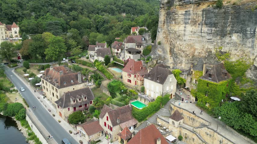 Drone aerial view in France countryside small old medieval town below a stone cliff surrounded by a green forest and a river circling around in La Roque Gageac