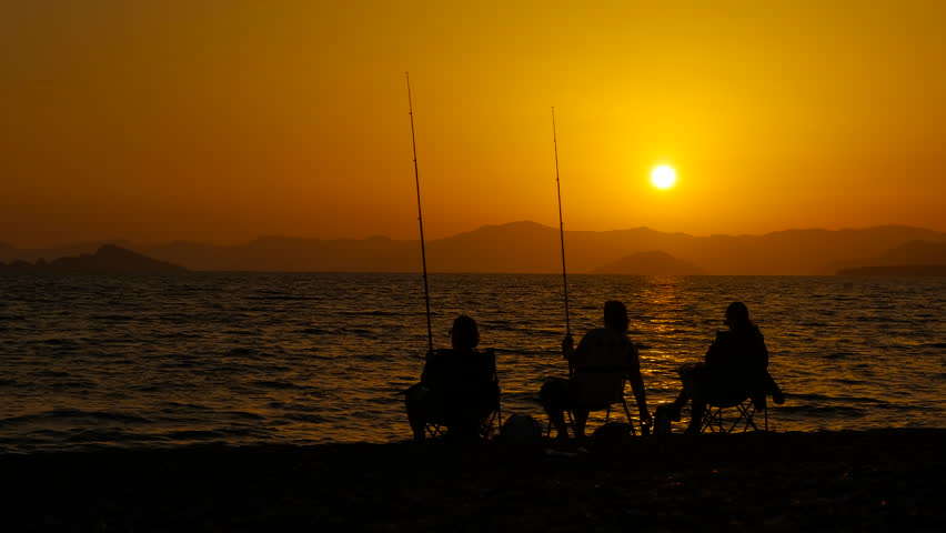 Silhouette of family leisure during fishing. A view of resting persons silhouette sitting in chairs on the beach and fishing during summer evening.