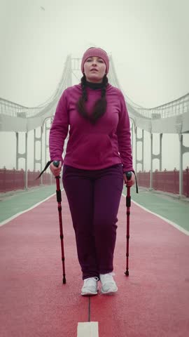Fit and beautiful chubby woman training with nordic walking poles, walking through bridge in fog. Confident motivated female in violet sportswear and winter headband walking outdoors. Vertical video