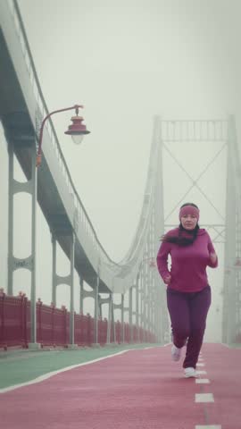 Motivated chubby female stops on bridge and hard breaths after intense running training in the foggy morning. Woman running outdoors, trying to lose weight, doing cardio workouts. Vertical video