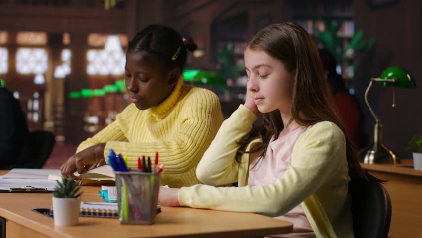 Preteen girls focus on reading the required books at a study desk, developing their academic skills and literary language at the library. Scholars enjoy lecture for the school curriculum. Camera B.