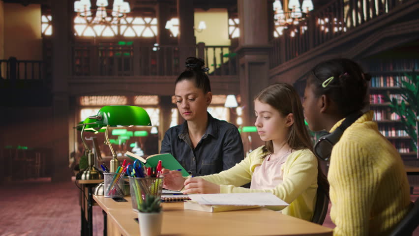 Female tutor mentoring schoolgirls in a school library setting, boosting their understanding of a subject through personalized guidance. Teacher doing private lessons with two pupils. Camera A.