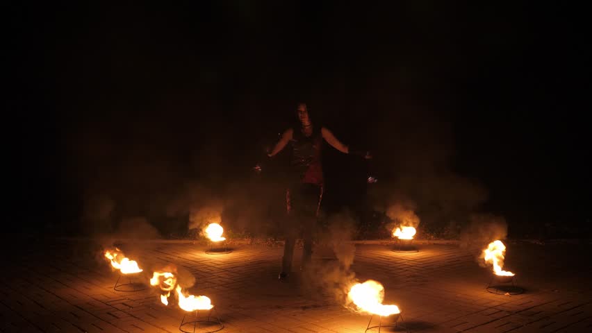 A woman fire-juggler, twirling long flaming cords in her hands. A nighttime circus act featuring fire. A pyrotechnic display. Fire-juggling. Slow motion.