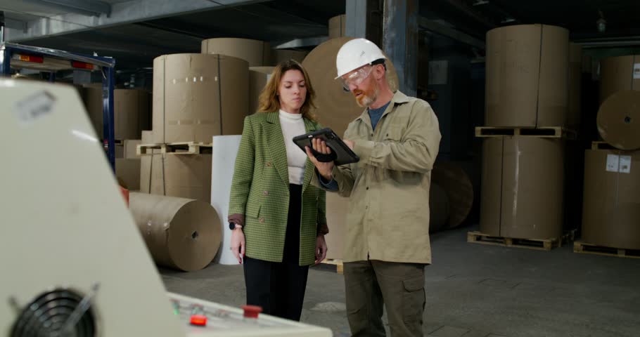 A worker and a boss talk and use a tablet while standing in the workshop at a paper mill.