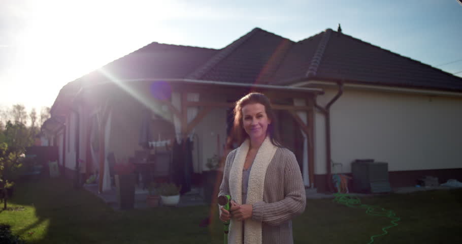 Woman holding garden hose in backyard, smiling near residential house, suburban home setting with patio, lawn, and greenery, casual outdoor gardening moment