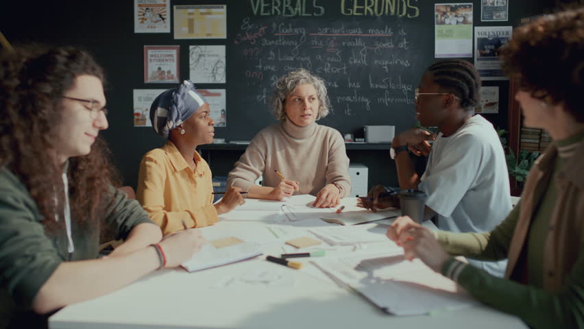 Female teacher and group of young international students sitting around table in classroom, discussing topics and taking notes in notebooks during English lesson