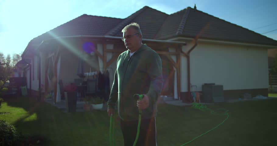 Man watering lawn with garden hose in backyard, casual moment with water spraying, residential house and greenery in suburban setting, relaxed and serene atmosphere