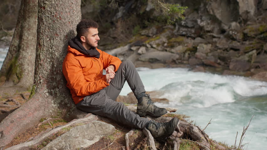 A young man sits comfortably against a tree by a river, enjoying the tranquility of the surrounding wilderness. He reflects peacefully as water flows nearby.