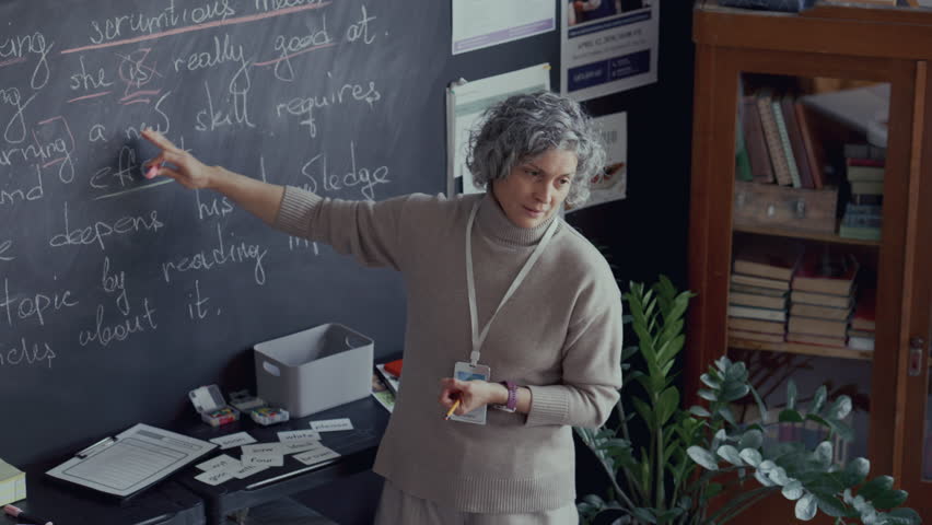 Mature female teacher with grey hair pointing at sentences written on chalkboard, explaining English grammar rules during class. High angle view