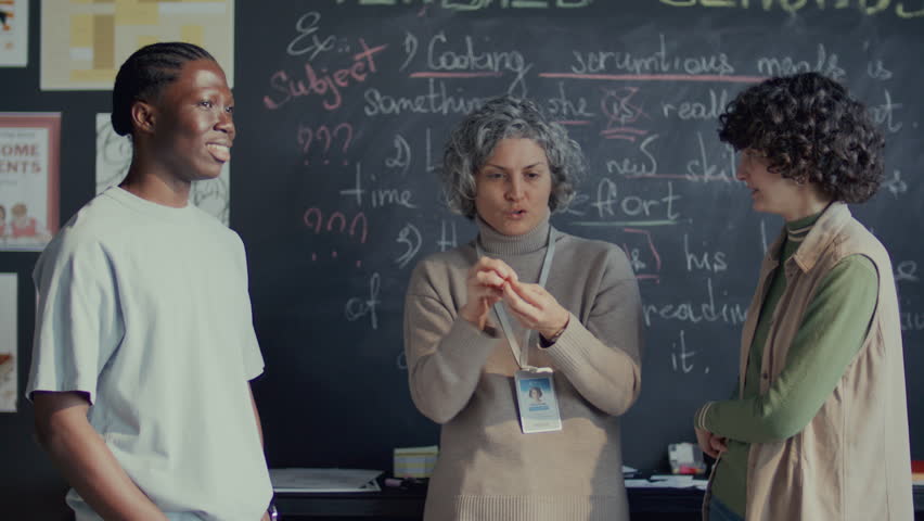 Middle-aged female teacher standing in front of blackboard in classroom, explaining some unclear points to young diverse students after English lesson