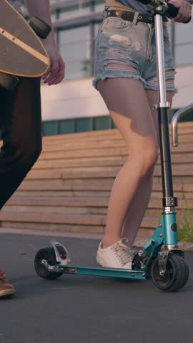 Pretty asian girl and handsome guy in front of orange sunset passing by architectural building or park with a board and scooter in hands. Friends communicating with each other while walking.
