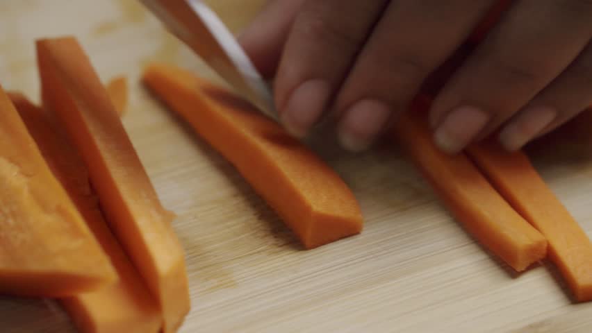Handheld shot of Preparing Fresh Carrot Sticks for Cooking by Slicing Them on a Wooden Board