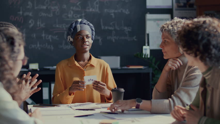 African American female student holding flashcard labeled Ate and discussing it with teacher and diverse classmates, learning new words during English lesson