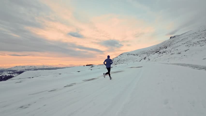 Young Caucasian male athlete runner walking in pashmina and warm clothes in winter landscape in mountains surrounded by snow and mountains at sunset. Jogging at dusk. Aerial view from fpv sports drone