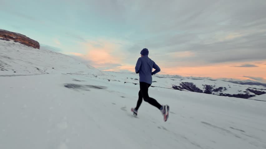 Young Caucasian male athlete runner walking in pashmina and warm clothes in winter landscape in mountains surrounded by snow and mountains at sunset. Jogging at dusk. Aerial view from fpv sports drone