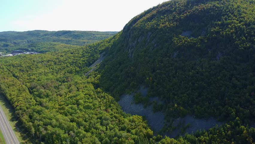 A panoramic aerial view showing the steep green cliffs of an old extinct volcano surrounded by forest and bordered by a country road. Atholville, New Brunswick, Canada, 2024.