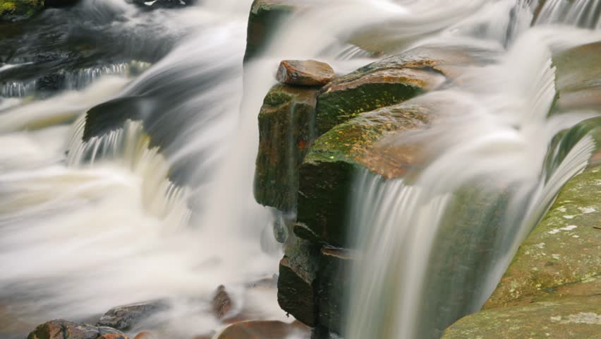 A long exposure shot of waterfall streaming on rocks with an old stone bridge in the background in Three Shires Head, Derbyshire, UK