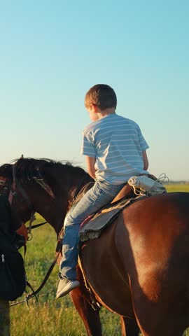 Father puts little boy on horse. Slow motion. Child riding horse under father supervision on farm. Child sitting on horse, walk in field. Happy family on farm. Dad teaching son to ride horse in saddle