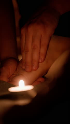 Masseur is giving a relaxing foot massage to a customer by candlelight in a spa. The masseuse is using their hands to massage the customer's feet