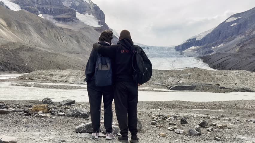 Man and a woman are standing in front of Athabasca Glacier. The man is wearing a black jacket and the woman is wearing a black hoodie. They are both holding backpacks, Canada