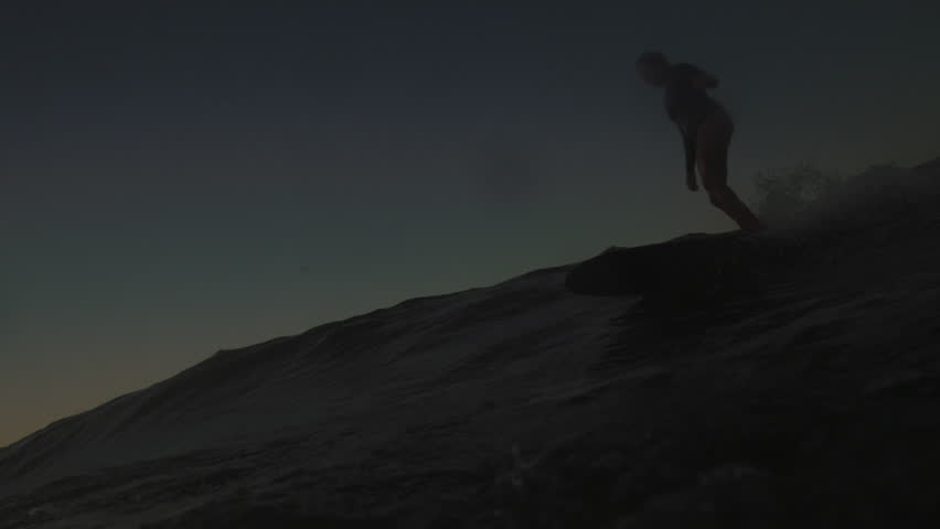 Tracking follows silhouette of female surfer walking on longboard and then falling off at sunset