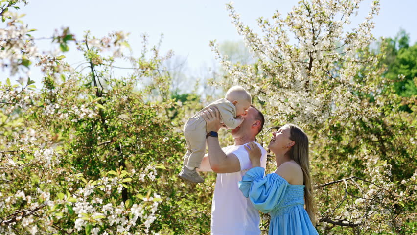 Caucasian dad tossing his adorable baby boy. Mother stands behind her husband smiling to a kid. Blossoming garden at backdrop.