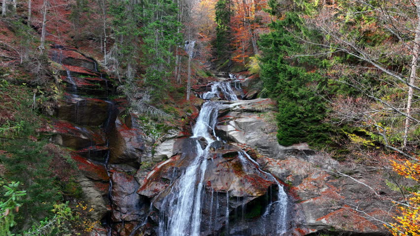 Tranquil forest scene with an autumn waterfall, golden leaves covering the ground, and silky water flowing over rugged stone formations. Aerial nature video.